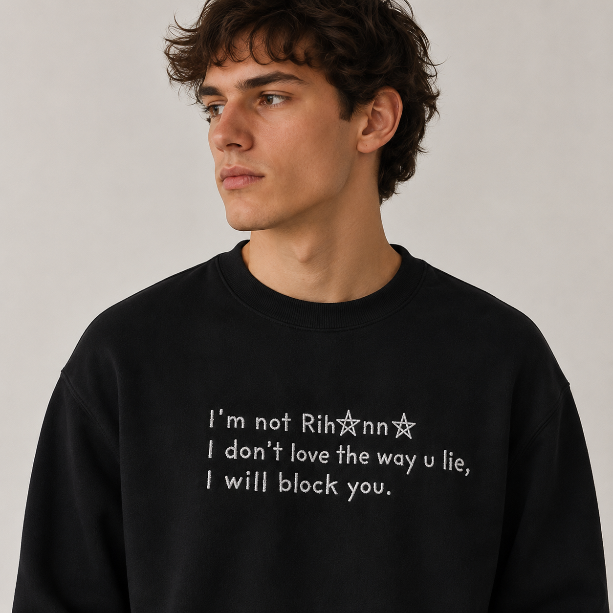 Young man wearing a black crewneck sweatshirt embroidered with white text reading "I'm not Rihanna I don't love the way u lie, I will block you."