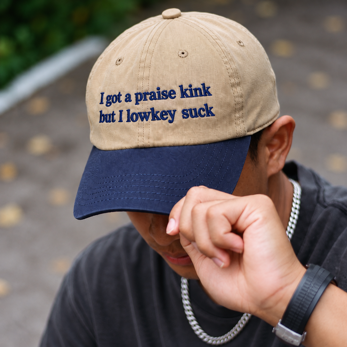 A man wearing a beige and navy blue baseball cap with the embroidered text 'I got a praise kink but I lowkey suck', photographed outdoors with his hand near his face.