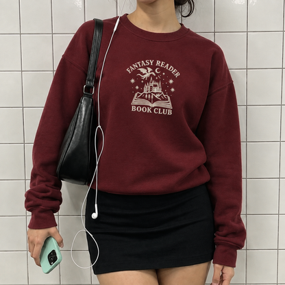 A person wearing a burgundy crewneck sweatshirt with a cream graphic that reads 'FANTASY READER BOOK CLUB' and an illustration of a castle and open book, paired with a black mini skirt and carrying a black shoulder bag.