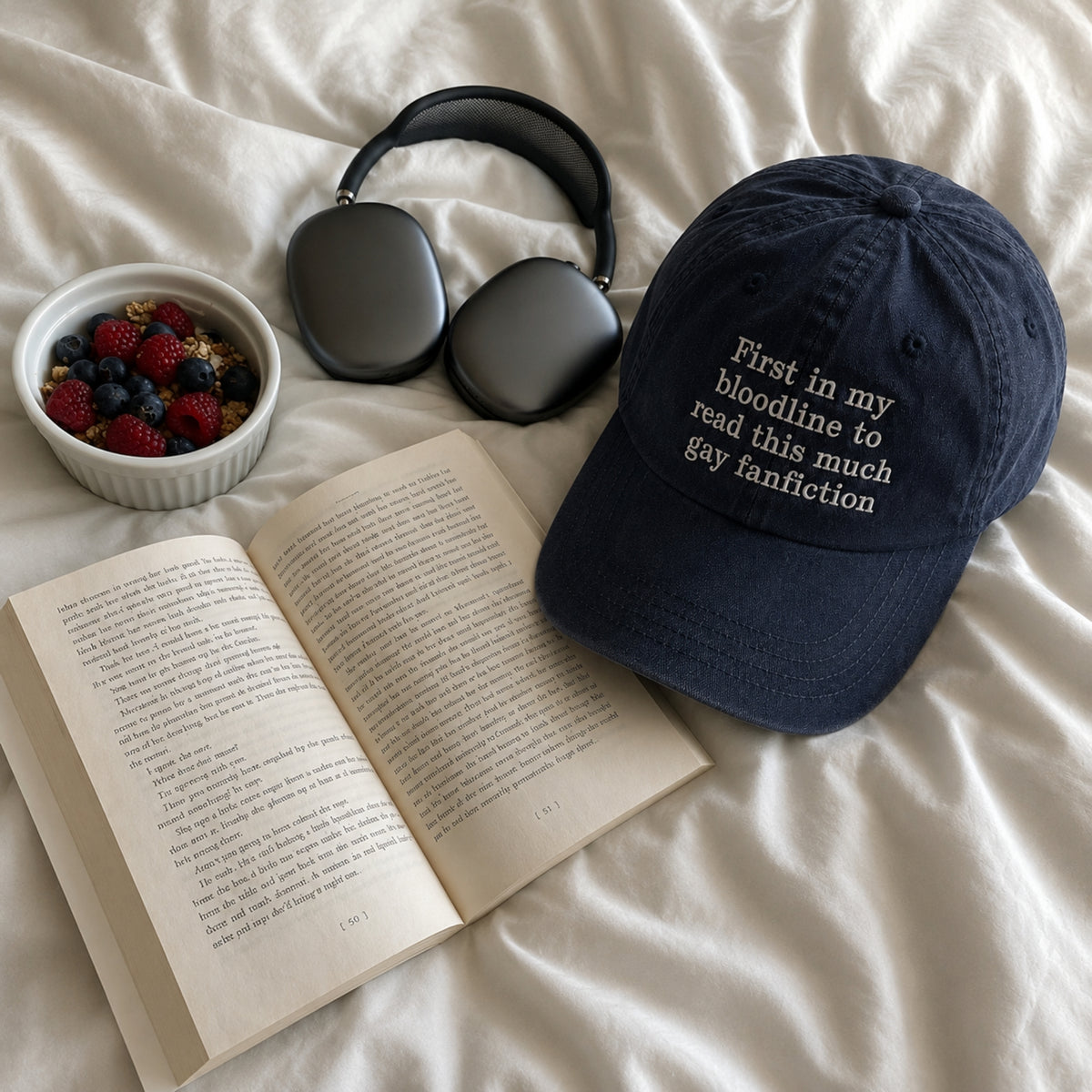 A navy blue baseball cap with white embroidered text reading 'First in my bloodline to read this much gay fanfiction' is styled on white bedding alongside an open book, a bowl of mixed berries, and dark gray over-ear headphones.
