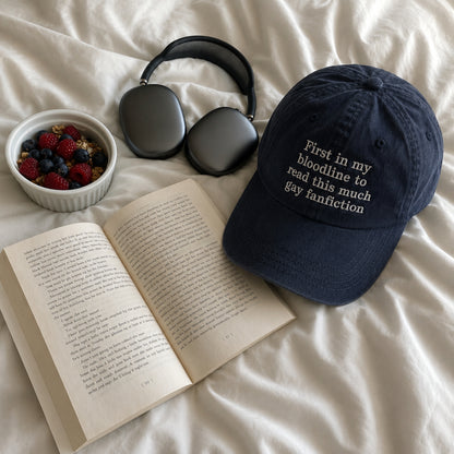 A navy blue baseball cap with white embroidered text reading 'First in my bloodline to read this much gay fanfiction' is styled on white bedding alongside an open book, a bowl of mixed berries, and dark gray over-ear headphones.