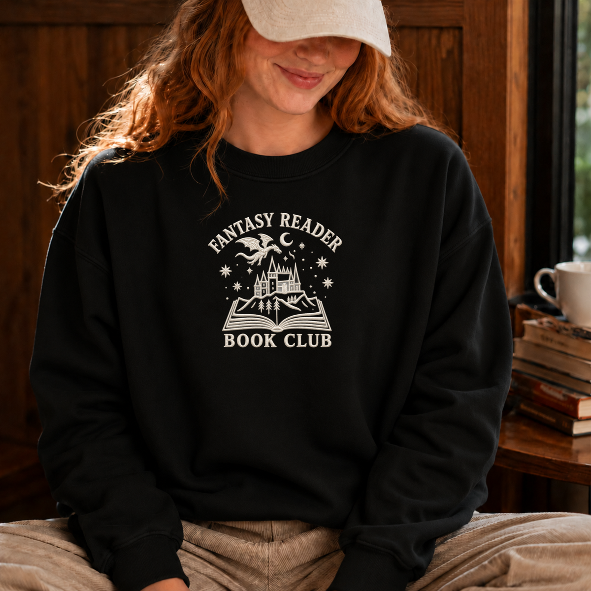 A woman wearing a black crewneck sweatshirt with a white graphic that reads 'FANTASY READER BOOK CLUB' and shows a cozy indoor setting with a wooden background.