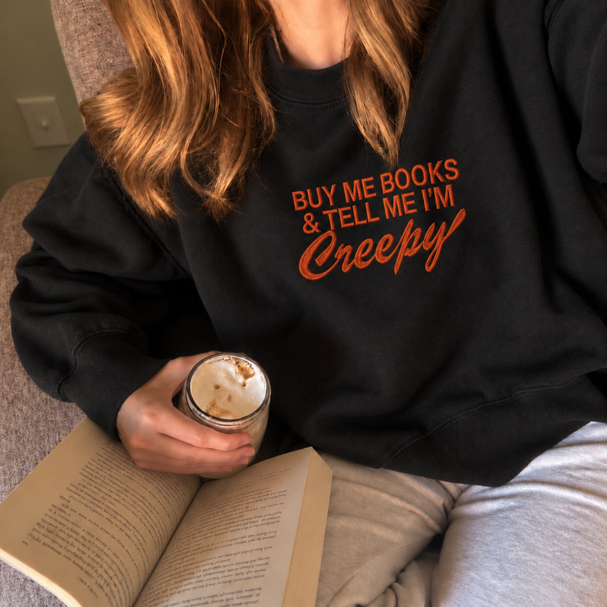 A person wearing a black crewneck sweatshirt with orange text that reads 'BUY ME BOOKS & TELL ME I'M Creepy', holding an open book and a drink while sitting indoors.