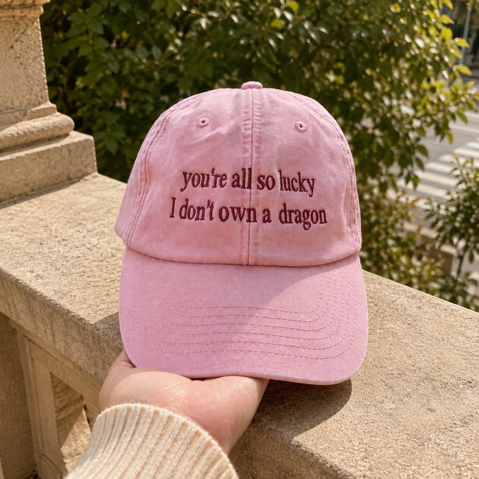 A pink baseball cap held in a hand, featuring the embroidered text 'you're all so lucky I don't own a dragon' in dark pink lettering. The background shows a stone wall and green foliage.