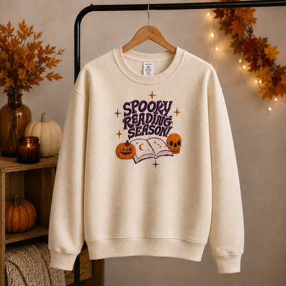 An off-white crewneck sweatshirt hanging on a wooden hanger from a black clothing rack, decorated with a 'Spooky Reading Season' graphic featuring a purple book, a jack-o'-lantern, and a skull. The background is a warm, cozy autumn scene with pumpkins, a vase of leaves, and string lights.