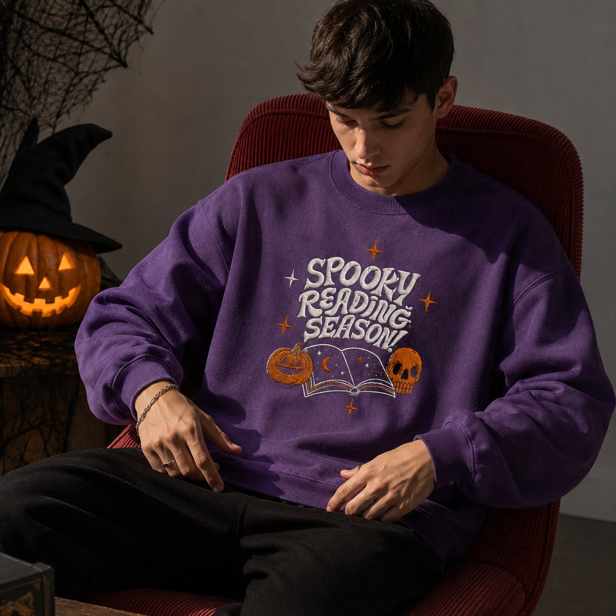 A man wearing a purple crewneck sweatshirt with a 'Spooky Reading Season!' graphic, sitting in a red chair against a gray wall.