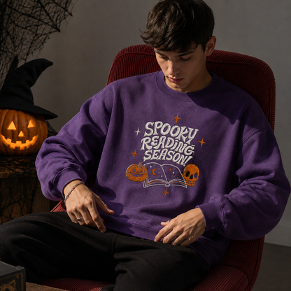 A man wearing a purple crewneck sweatshirt with a 'Spooky Reading Season!' graphic, sitting in a red chair against a gray wall.