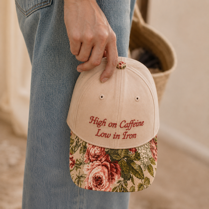 Hand holding a beige baseball cap with red embroidered text 'High on Caffeine Low in Iron' and a vintage floral rose tapestry visor against blue jeans.