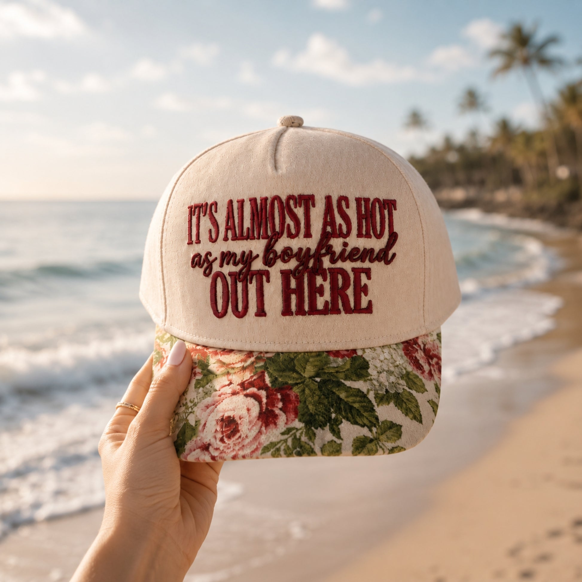 A hand holds a beige baseball cap with dark red embroidered text reading 'IT'S ALMOST AS HOT as my boyfriend OUT HERE' above a floral brim, set against a blurred beach background with ocean waves and palm trees.