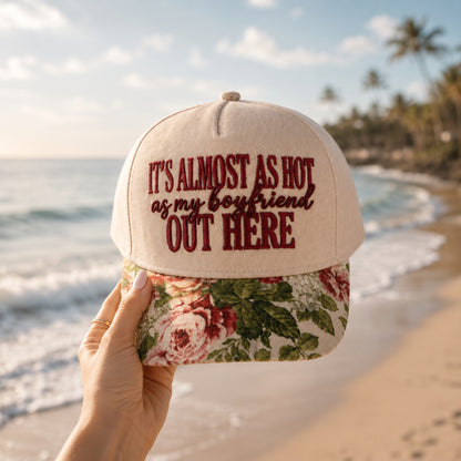 A hand holds a beige baseball cap with dark red embroidered text reading 'IT'S ALMOST AS HOT as my boyfriend OUT HERE' above a floral brim, set against a blurred beach background with ocean waves and palm trees.