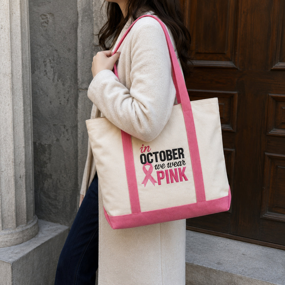 A woman wearing a cream cardigan and navy pants carries a cream canvas tote bag with pink trim and straps. The bag features the printed text 'in OCTOBER we wear PINK' with a pink ribbon graphic.