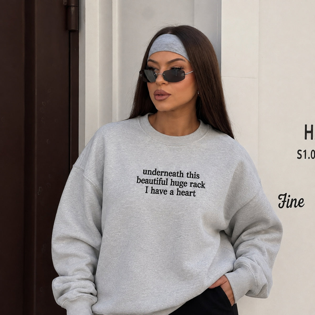 A woman wearing a light gray oversized crewneck sweatshirt with black text on the chest, styled with a gray headband and sunglasses.