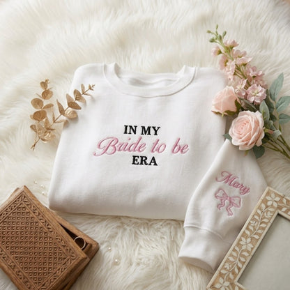 A white crewneck sweatshirt with 'IN MY Bride to be ERA' embroidered on the chest and 'Mary' with a bow on the sleeve, styled in a flat lay on a white faux fur background with pink roses, a brown woven box, and a wooden frame.