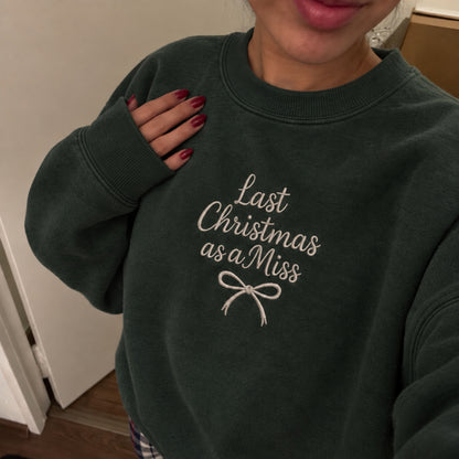 A person wearing a dark forest green crewneck sweatshirt with the text 'Last Christmas as a Miss' and a bow graphic embroidered in white on the front.