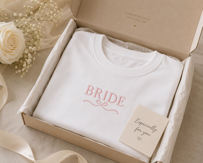 A white sweatshirt with pink "BRIDE" embroidery displayed inside an open brown cardboard box, accompanied by a small "Especially for you" card with a heart, a white rose, greenery, and a beige ribbon.