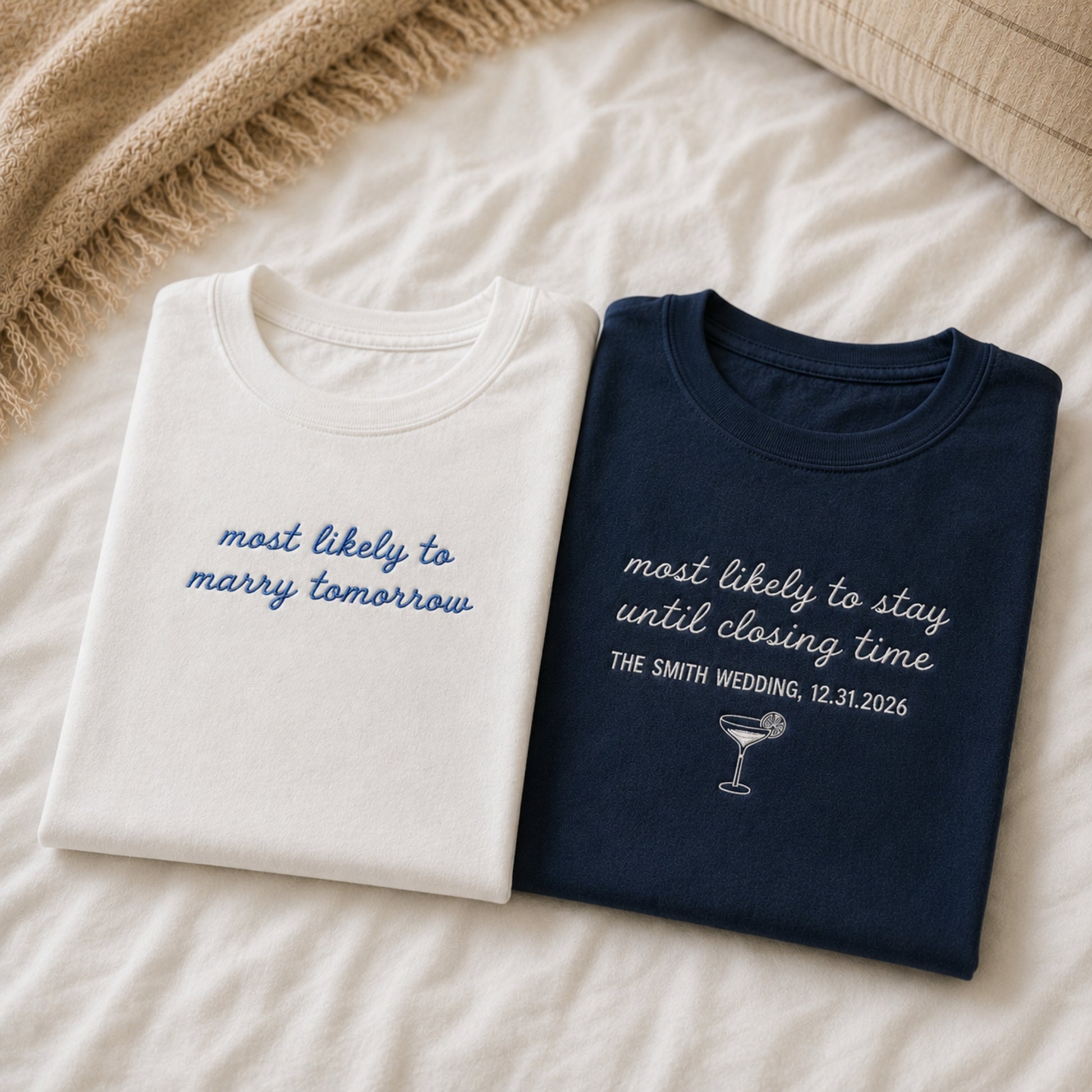 Two folded graphic T-shirts laid flat on a cream bedspread, with a beige knitted throw at the top left. The left shirt is off-white with blue script text reading 'most likely to marry tomorrow.