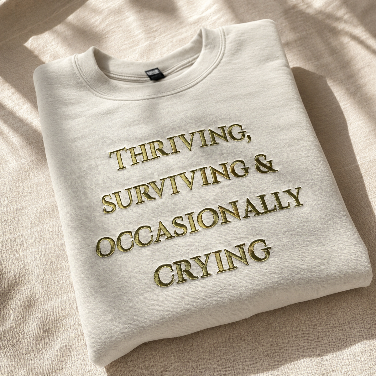 A folded off-white crewneck sweatshirt with gold embroidered text reading 'THRIVING, SURVIVING & OCCASIONALLY CRYING' on a textured beige fabric background with soft leaf shadows.