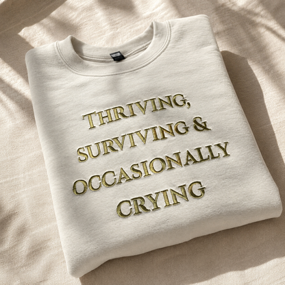 A folded off-white crewneck sweatshirt with gold embroidered text reading 'THRIVING, SURVIVING & OCCASIONALLY CRYING' on a textured beige fabric background with soft leaf shadows.