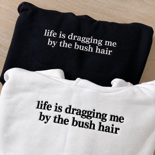 Two folded graphic t-shirts on a light wood background. The top shirt is black with white text reading 'life is dragging me by the bush hair', and the bottom shirt is white with black text reading the same phrase.