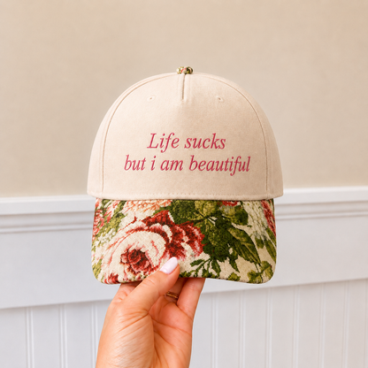 A hand holds a beige baseball cap with red embroidered text reading 'Life sucks but i am beautifull' and a floral patterned visor in red, green, and cream tones, against a neutral indoor background.
