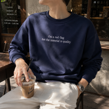 Person sitting at an outdoor cafe holding an iced coffee, wearing white pants and a navy blue crewneck sweatshirt embroidered with white text reading "i'm a red flag but the material is quality".