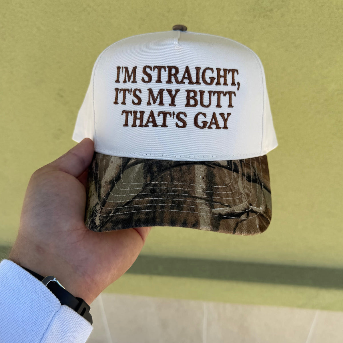 A hand holding a white and camouflage trucker hat with the text 'I'M STRAIGHT, IT'S MY BUTT THAT'S GAY' printed in brown on the front, set against a textured yellow wall.