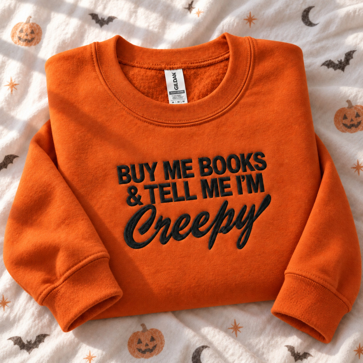 An orange crewneck sweatshirt with black embroidered text reading 'BUY ME BOOKS & TELL ME I'M Creepy' is laid flat on a white fabric background with a Halloween pattern of pumpkins, bats, and moons. The Gildan brand tag is visible inside the collar.