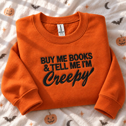 An orange crewneck sweatshirt with black embroidered text reading 'BUY ME BOOKS & TELL ME I'M Creepy' is laid flat on a white fabric background with a Halloween pattern of pumpkins, bats, and moons. The Gildan brand tag is visible inside the collar.