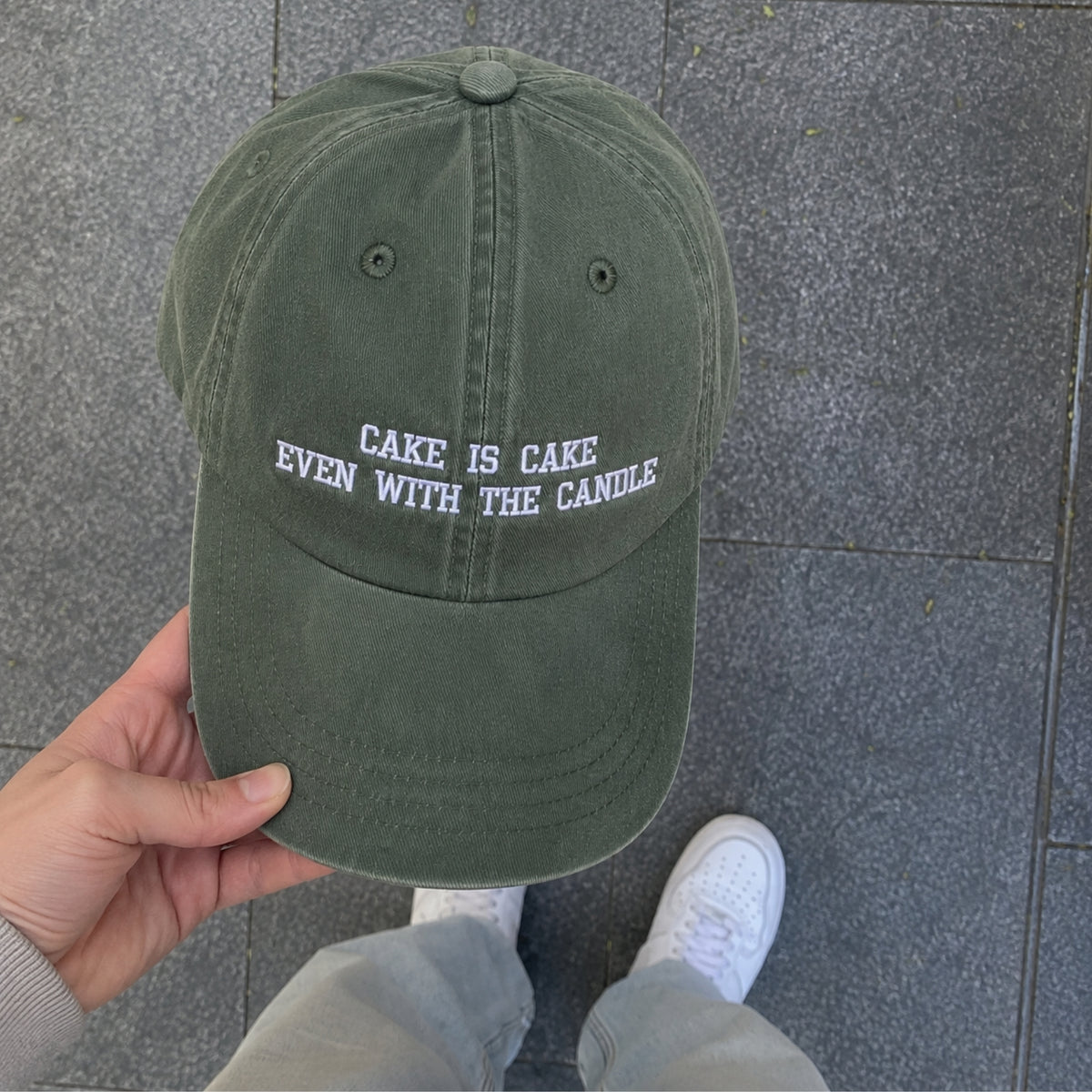An olive green dad hat with white embroidered text reading 'CAKE IS CAKE EVEN WITH THE CANDLE' is held by a hand over a gray tiled floor. The person's jeans and white sneakers are visible at the bottom.