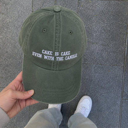 An olive green dad hat with white embroidered text reading 'CAKE IS CAKE EVEN WITH THE CANDLE' is held by a hand over a gray tiled floor. The person's jeans and white sneakers are visible at the bottom.