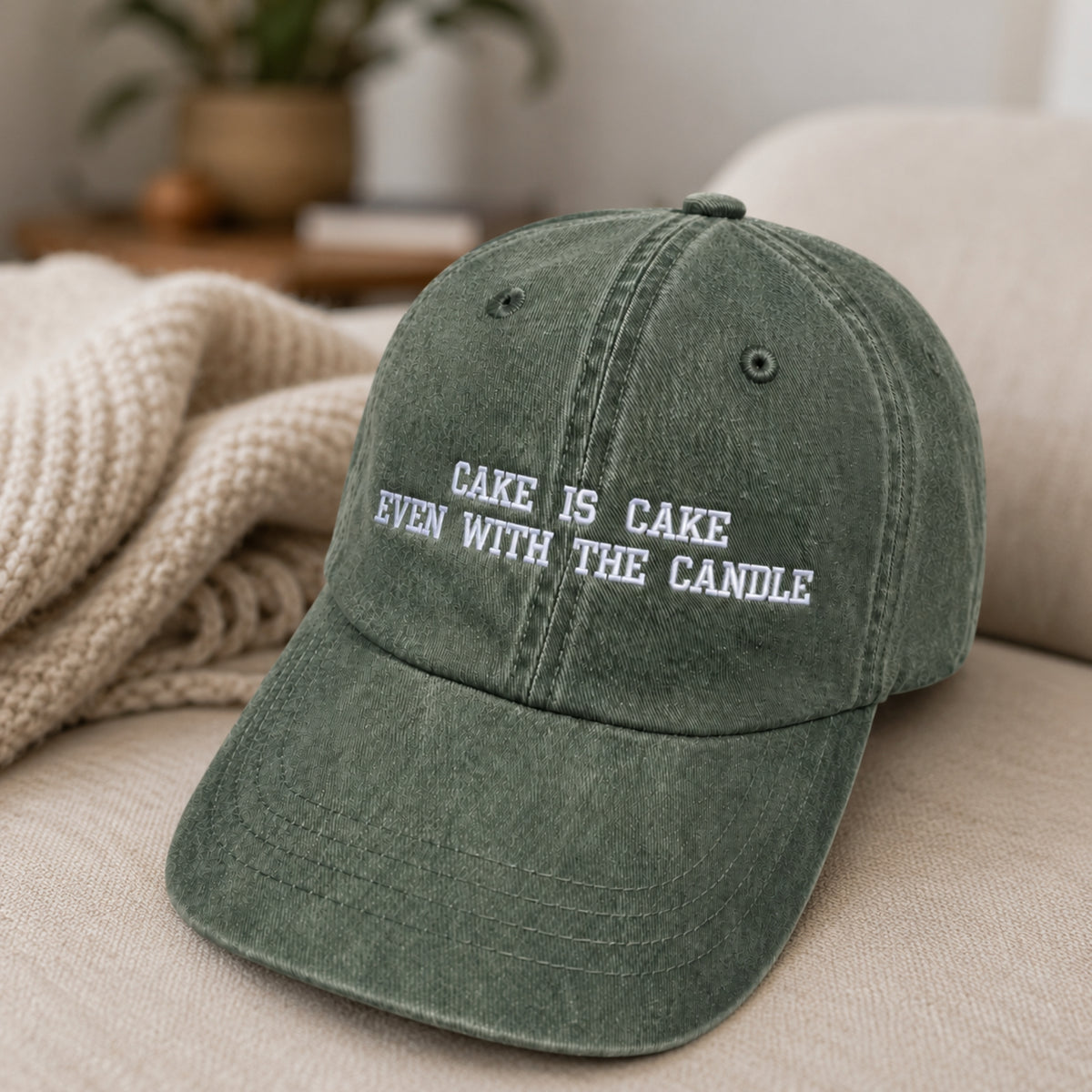 A washed olive green baseball cap with white embroidered text reading 'CAKE IS CAKE EVEN WITH THE CANDLE', resting on a beige sofa with a knitted blanket in the background.