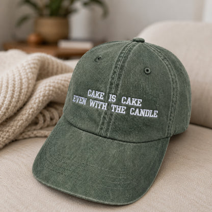 A washed olive green baseball cap with white embroidered text reading 'CAKE IS CAKE EVEN WITH THE CANDLE', resting on a beige sofa with a knitted blanket in the background.
