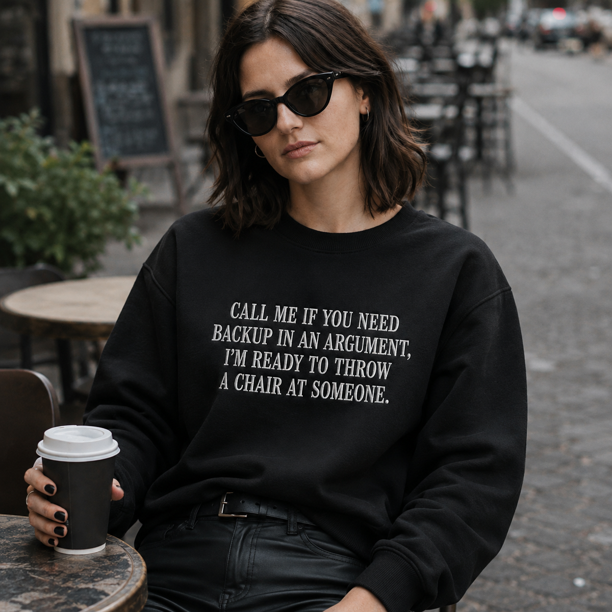 A woman wearing a black crewneck sweatshirt with white text that reads 'CALL ME IF YOU NEED BACKUP IN AN ARGUMENT, I'M READY TO THROW A CHAIR AT SOMEONE.' sits at an outdoor cafe table holding a coffee cup.