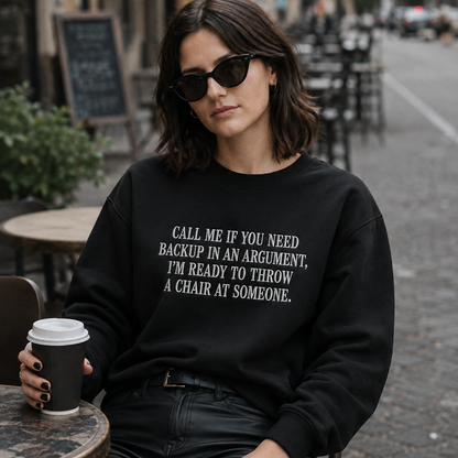 A woman wearing a black crewneck sweatshirt with white text that reads 'CALL ME IF YOU NEED BACKUP IN AN ARGUMENT, I'M READY TO THROW A CHAIR AT SOMEONE.' sits at an outdoor cafe table holding a coffee cup.