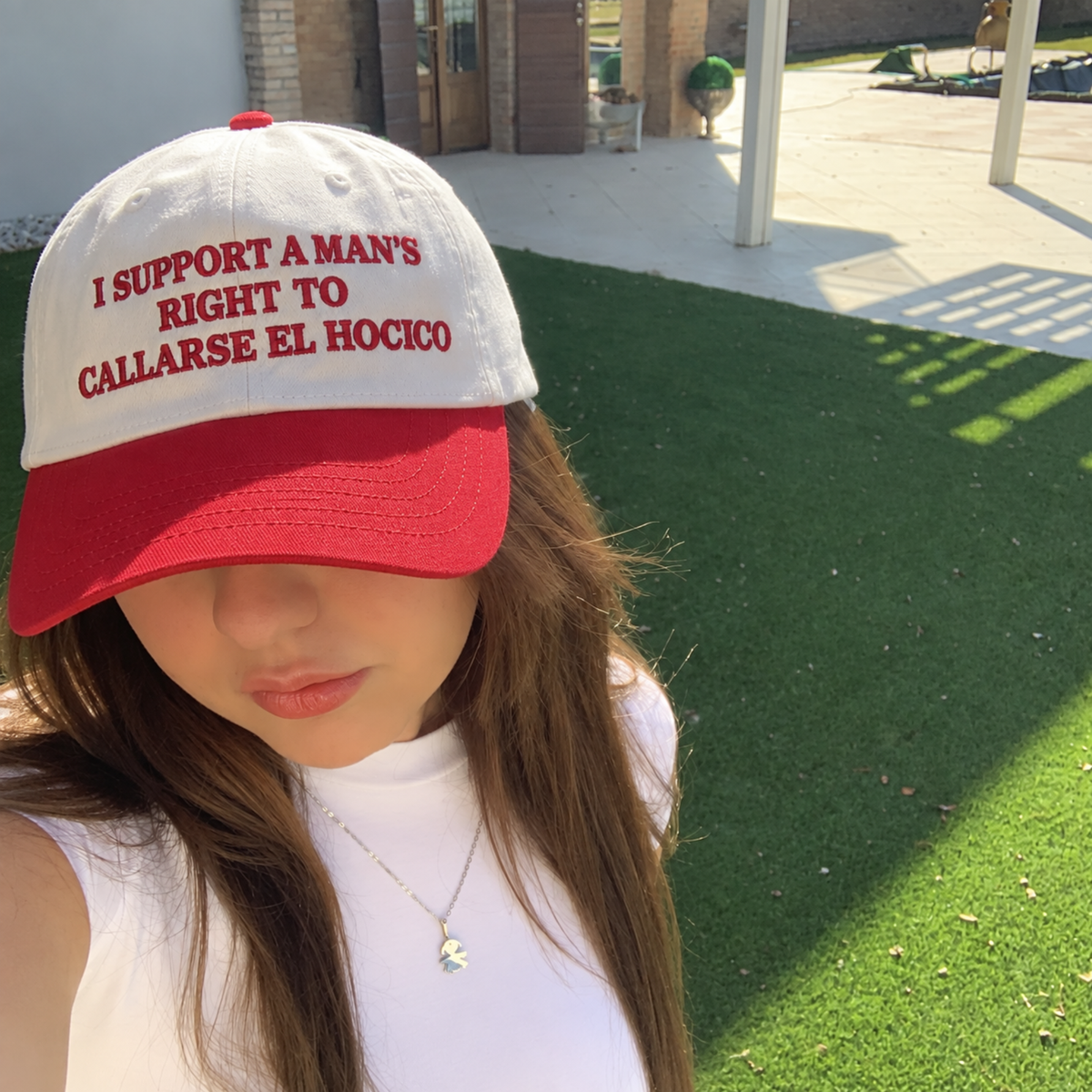 A person outdoors on a sunny day wearing a white baseball cap with a red brim. The cap has red embroidered text that reads, 'I SUPPORT A MAN'S RIGHT TO CALLARSE EL HOCICO'.
