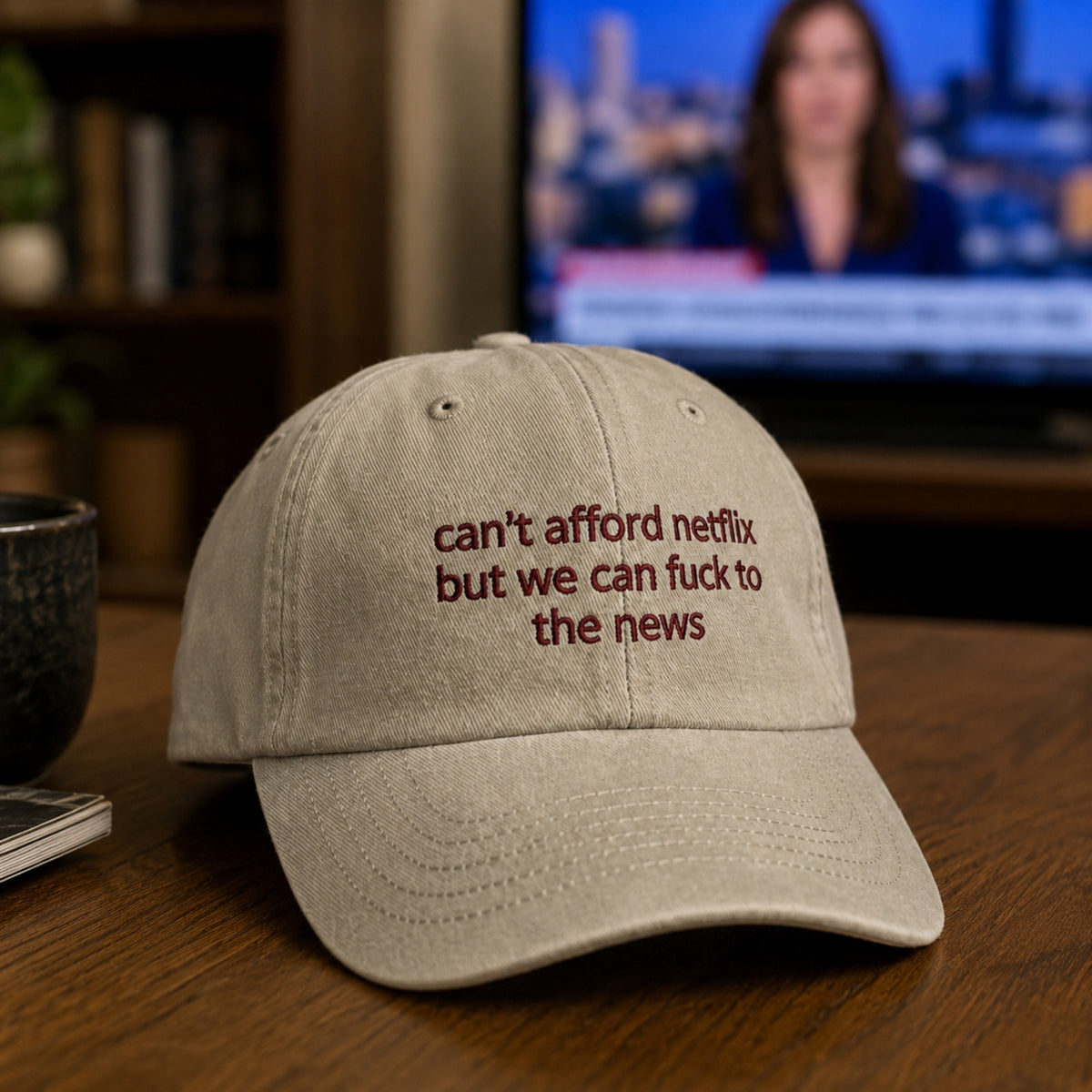 A light beige baseball cap with dark red embroidered text reading 'can't afford netflix but we can fuck to the news' on a wooden table, with a blurred TV and room background.