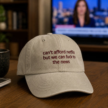 A light beige baseball cap with dark red embroidered text reading 'can't afford netflix but we can fuck to the news' on a wooden table, with a blurred TV and room background.