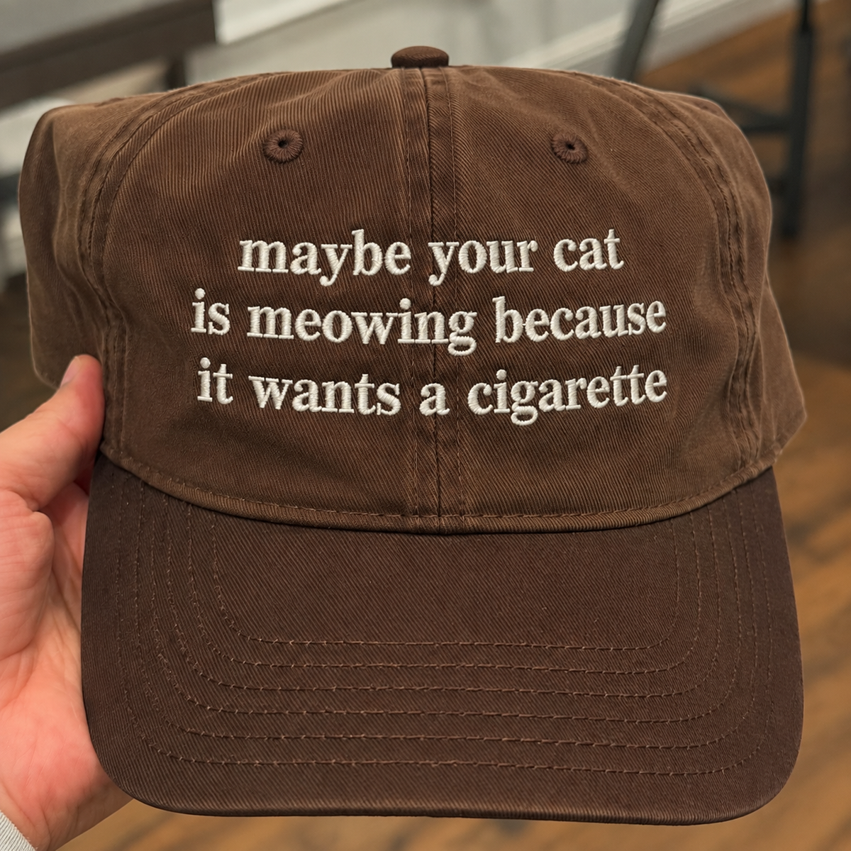 A brown cotton baseball cap held by a hand, featuring white embroidered text that reads 'maybe your cat is meowing because it wants a cigarette'.