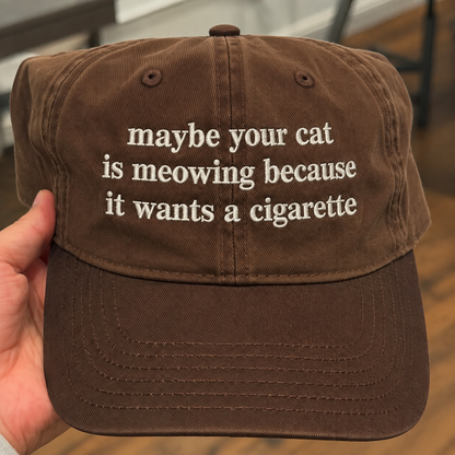 A brown cotton baseball cap held by a hand, featuring white embroidered text that reads 'maybe your cat is meowing because it wants a cigarette'.