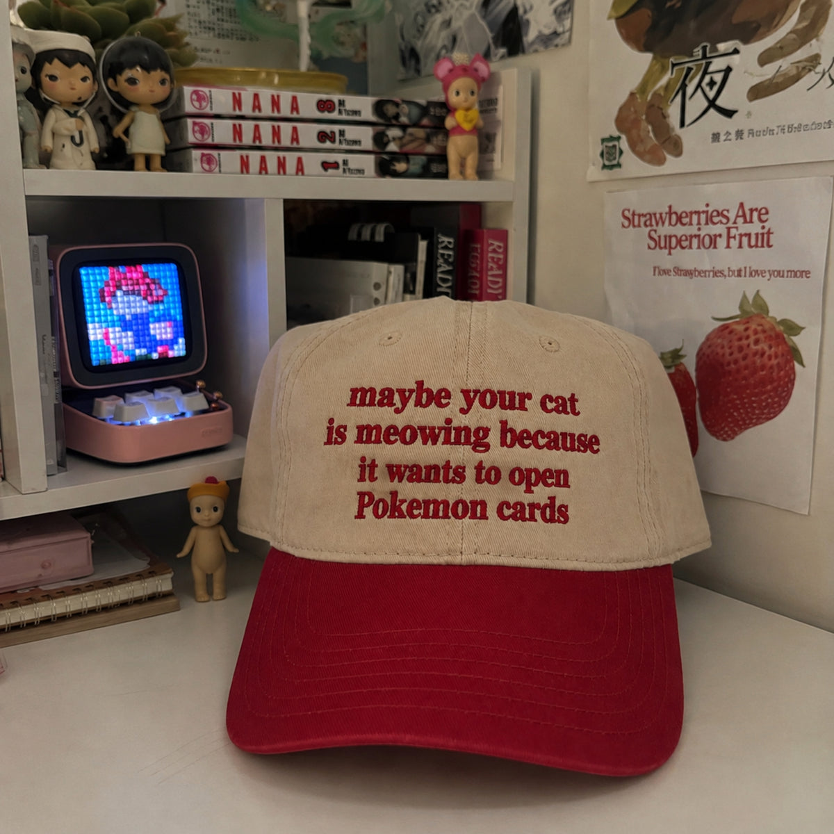 A baseball cap with a beige crown and a red brim, featuring red embroidered text that reads 'maybe your cat is meowing because it wants to open Pokemon cards'. The cap is displayed on a white desk in a room setting with bookshelves, a pixel art lamp, and a strawberry poster in the background.