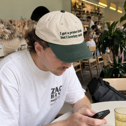 A man sitting at a cafe table wearing a beige and dark green baseball cap with the text 'I got a praise kink but I lowkey suck' and a white t-shirt, looking down at a phone.