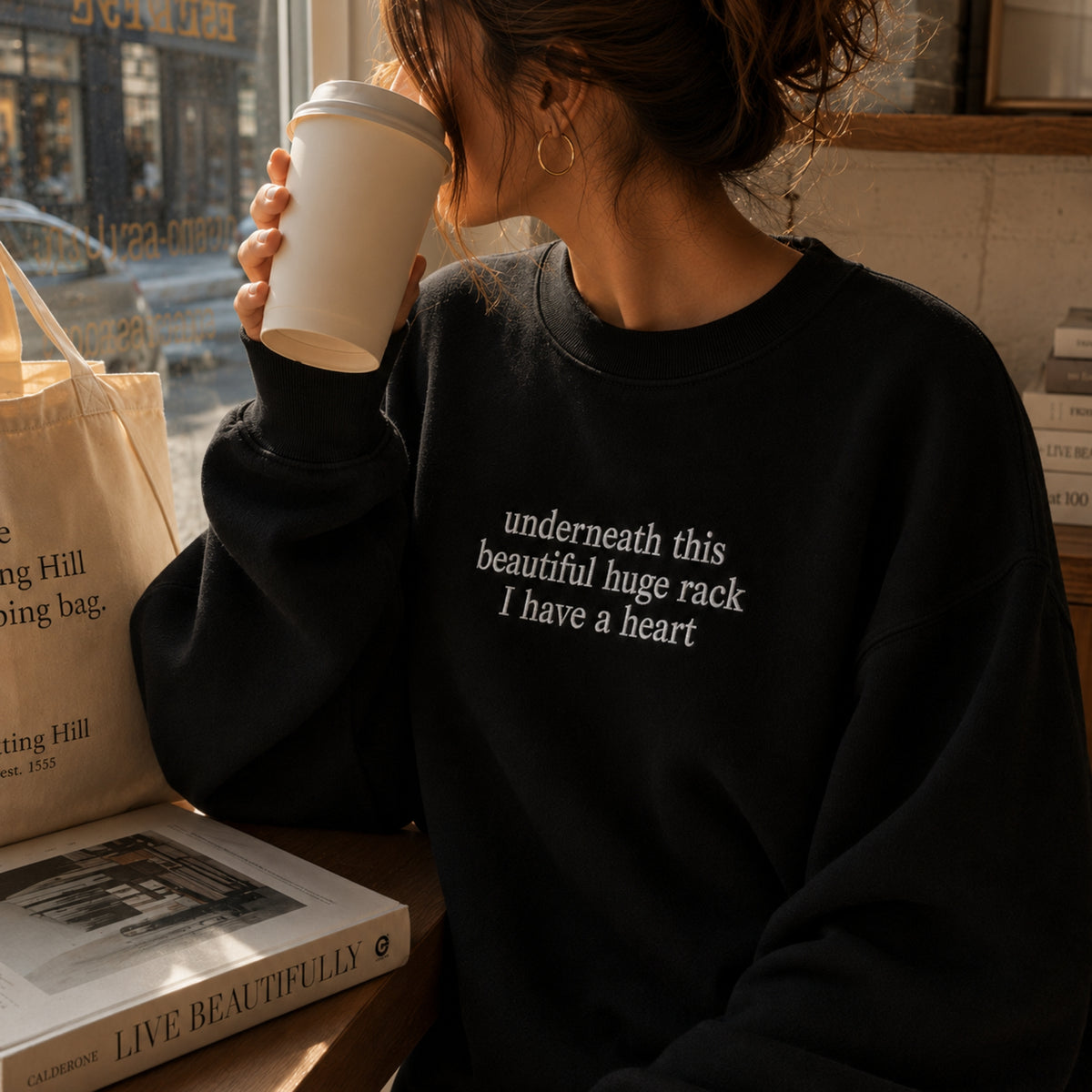 A woman in a cozy cafe setting wearing a black crewneck sweatshirt with white text that reads 'underneath this beautiful huge rack I have a heart'. She is holding a white paper coffee cup next to a canvas tote bag and a book on a wooden table.