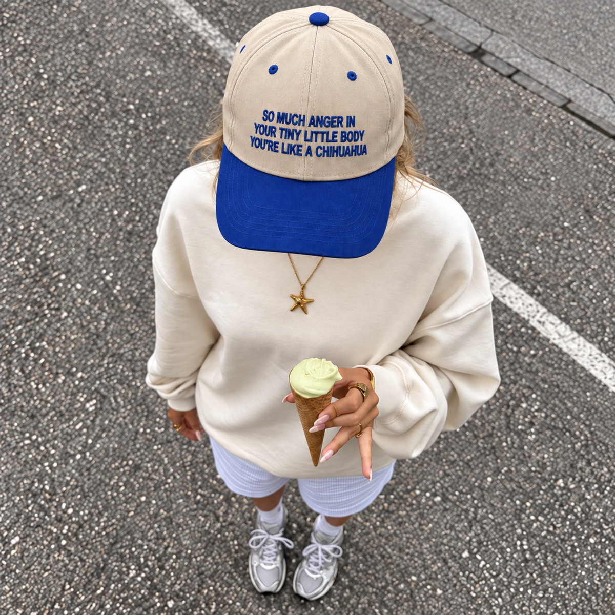 A person outdoors on asphalt wearing a cream sweatshirt, white shorts, gray sneakers, a blue and cream baseball cap with blue text, and holding an ice cream cone.