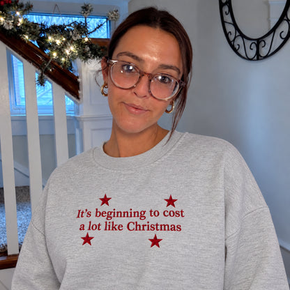 A woman wearing a light gray crewneck sweatshirt with red text reading 'It's beginning to cost a lot like Christmas' and red stars, standing indoors near a staircase with holiday decorations.