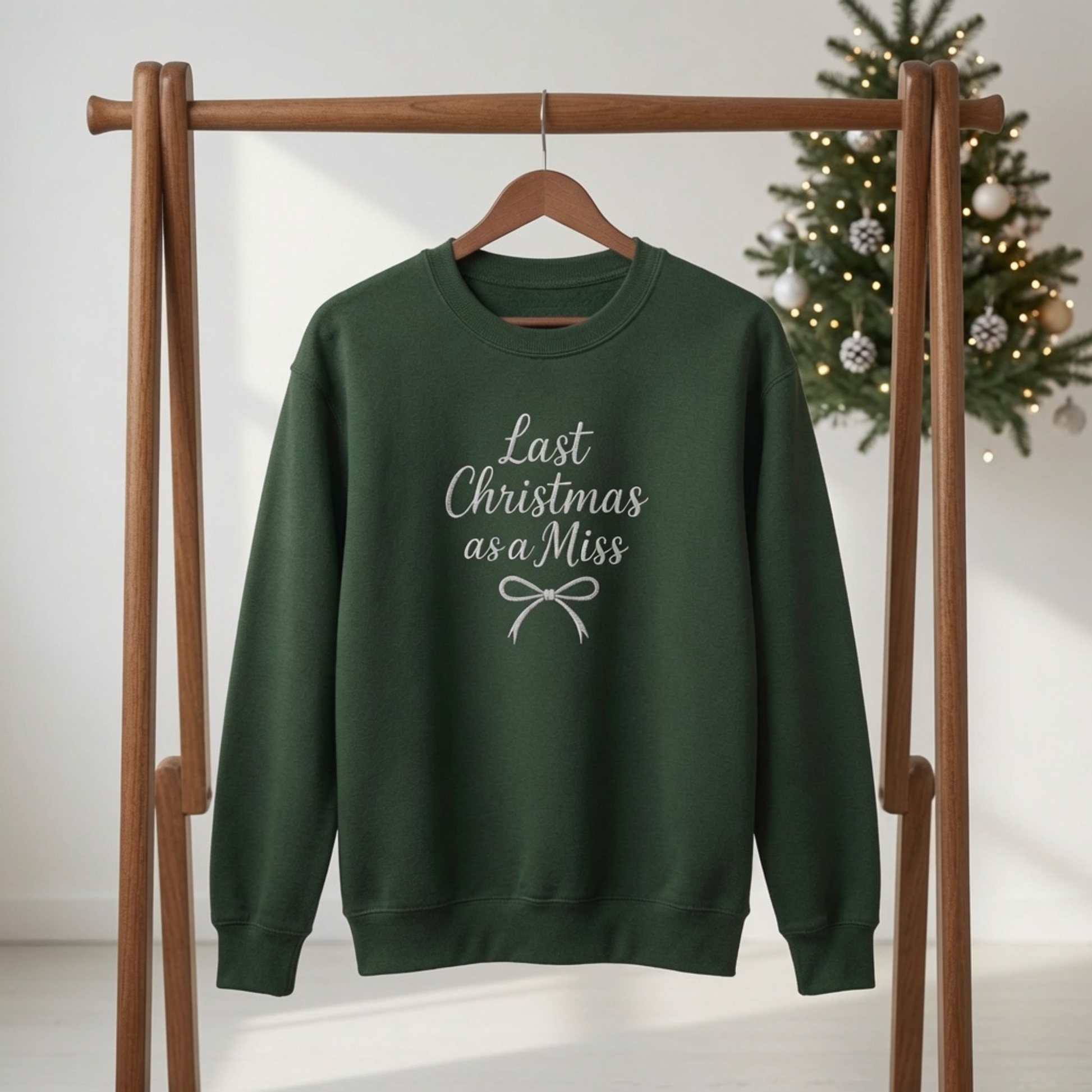 Forest green crewneck sweatshirt embroidered with white text reading "Last Christmas as a Miss" and a ribbon bow, hanging on a wooden rack in front of a Christmas tree.