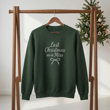 Forest green crewneck sweatshirt embroidered with white text reading "Last Christmas as a Miss" and a ribbon bow, hanging on a wooden rack in front of a Christmas tree.