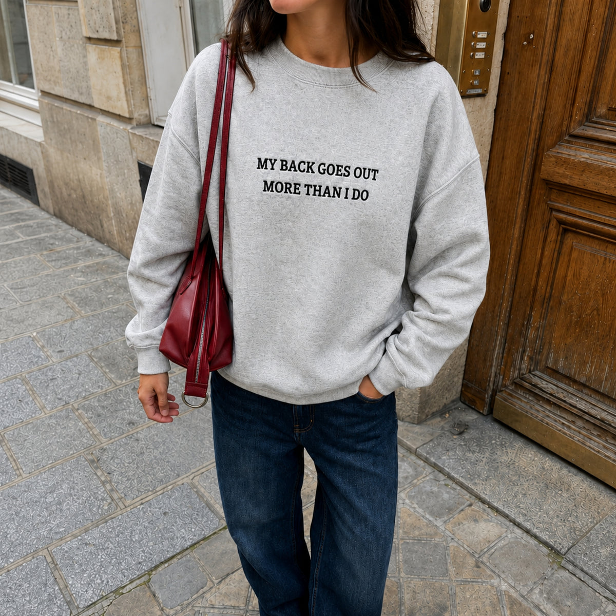 A woman wearing a light gray heather crewneck sweatshirt with the black printed text 'MY BACK GOES OUT MORE THAN I DO', paired with dark blue jeans and a dark red shoulder bag, standing on a stone sidewalk.