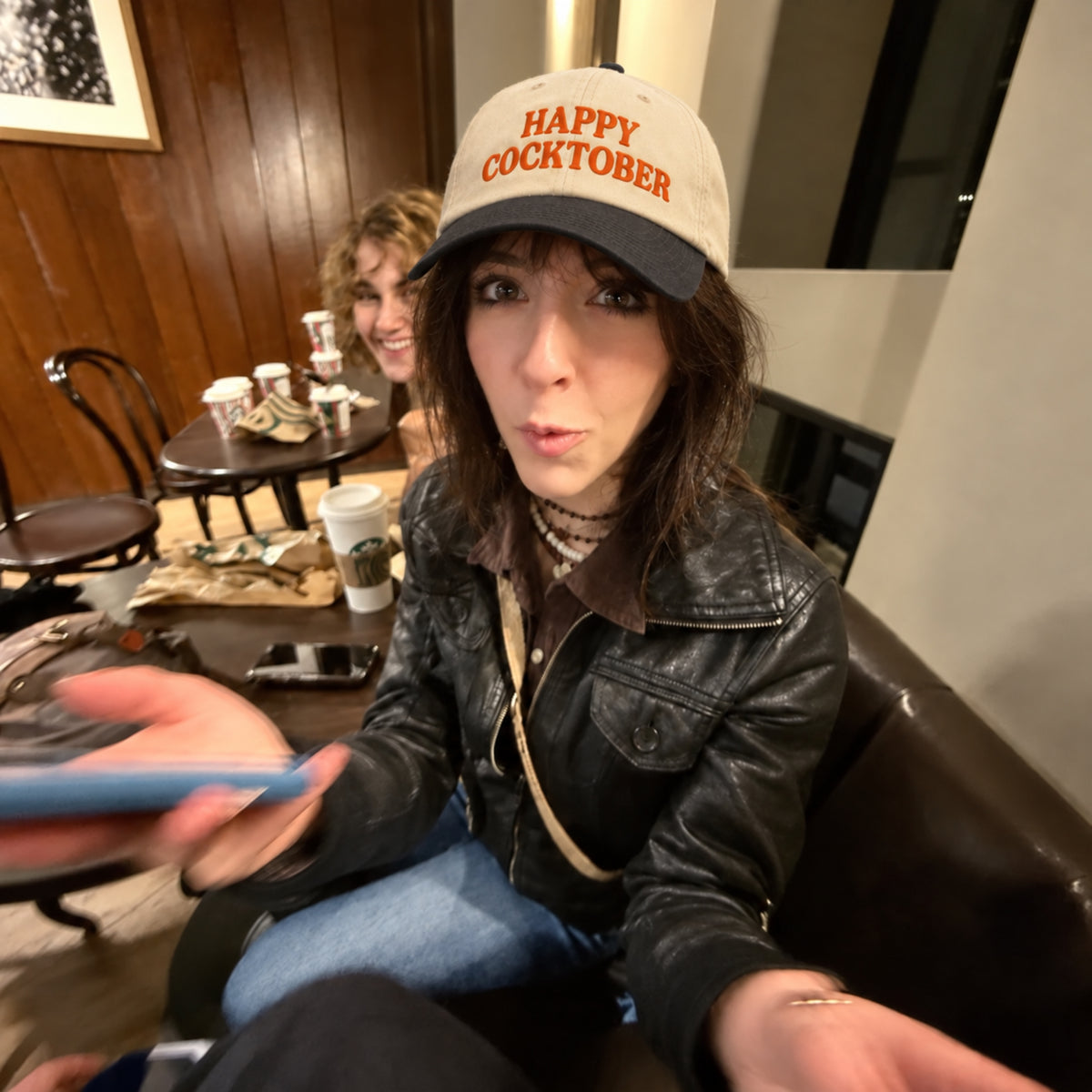 A woman wearing a beige and black trucker hat with 'HAPPY COCKTOBER' printed in orange, sitting in a dimly lit indoor setting with wood-paneled walls and a table in the background.
