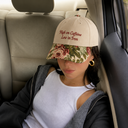 Woman sitting in a car wearing a beige baseball cap with red embroidered text 'High on Caffeine Low in Iron' and a vintage floral rose tapestry visor.