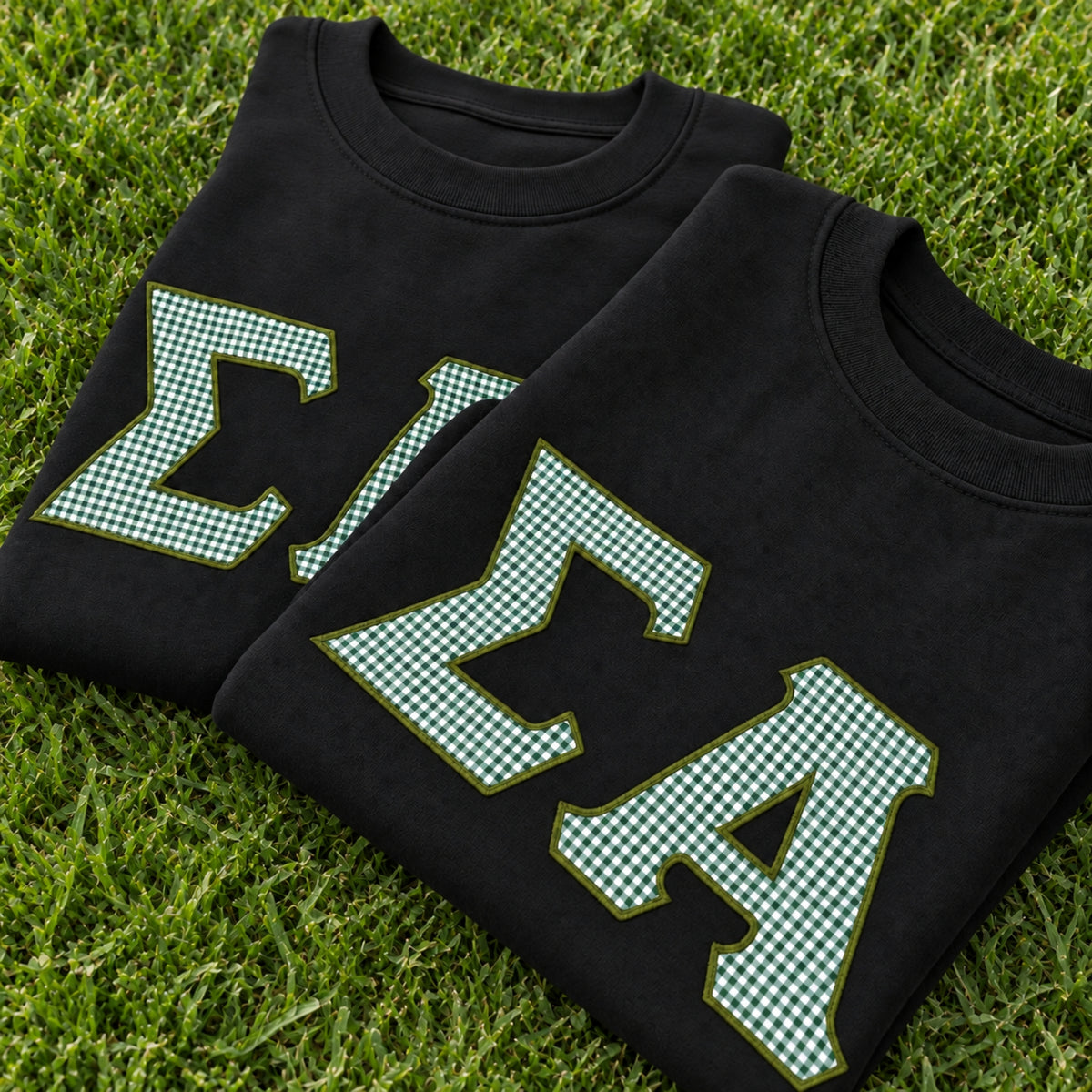 Two folded black T-shirts laid on green grass, each featuring large green-and-white gingham check letters outlined in olive green. The shirts are arranged diagonally, with one partially covering the other.